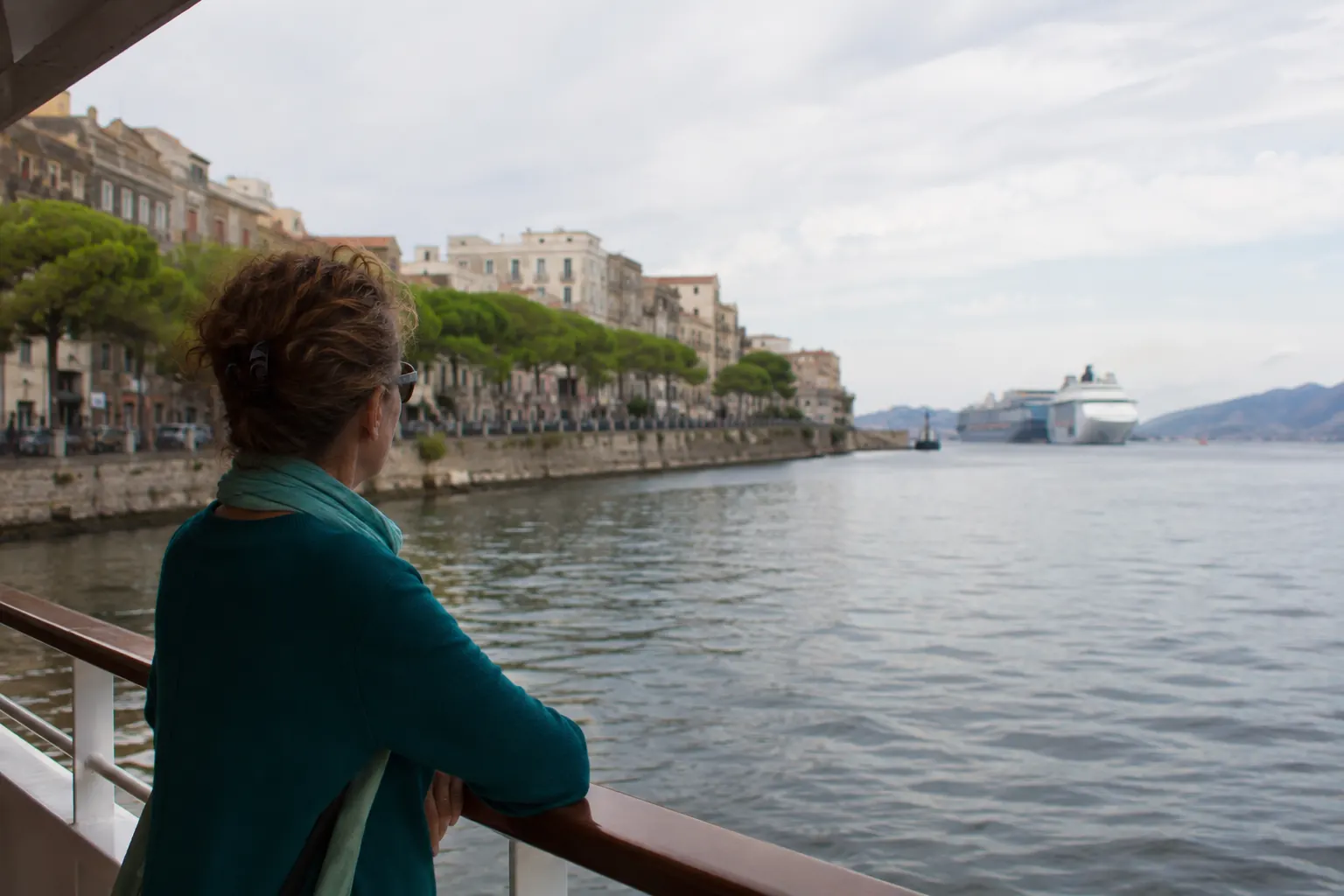 Traveler on a calm river-cruise deck with a distant ocean cruise ship beyond the river mouth.