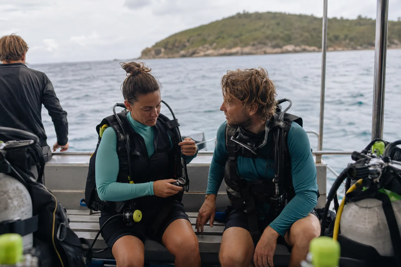 Scuba diver seated near the center of a dive boat, checking gear with a buddy while facing the horizon.