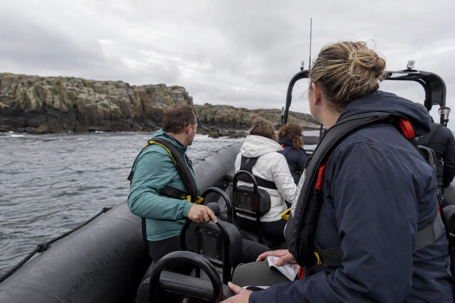 Life-jacketed passengers seated safely on a rigid inflatable boat idling near a rocky coast.