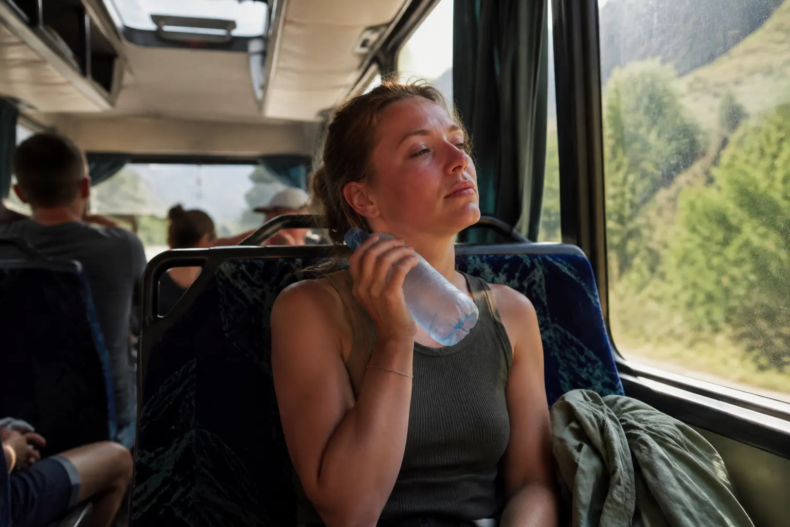 Warm bus passenger cooling their neck with a water bottle near an open ventilation window.