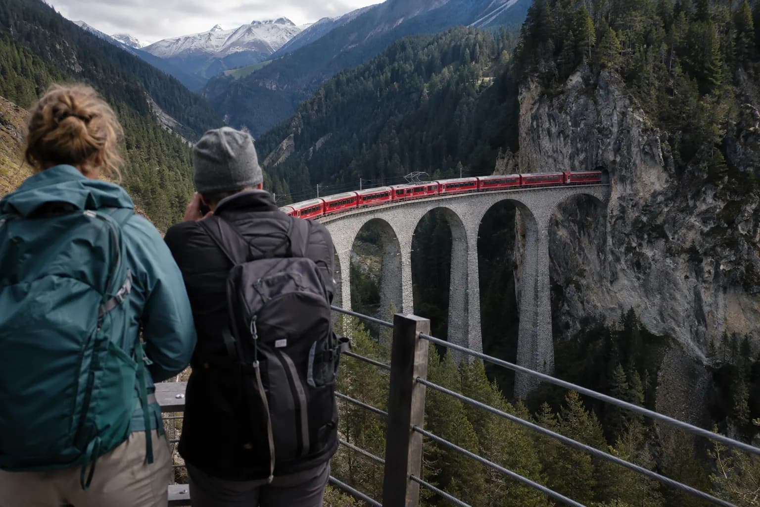 Mountain train rounding a stone viaduct in the Swiss Alps as hikers watch from a safe overlook.
