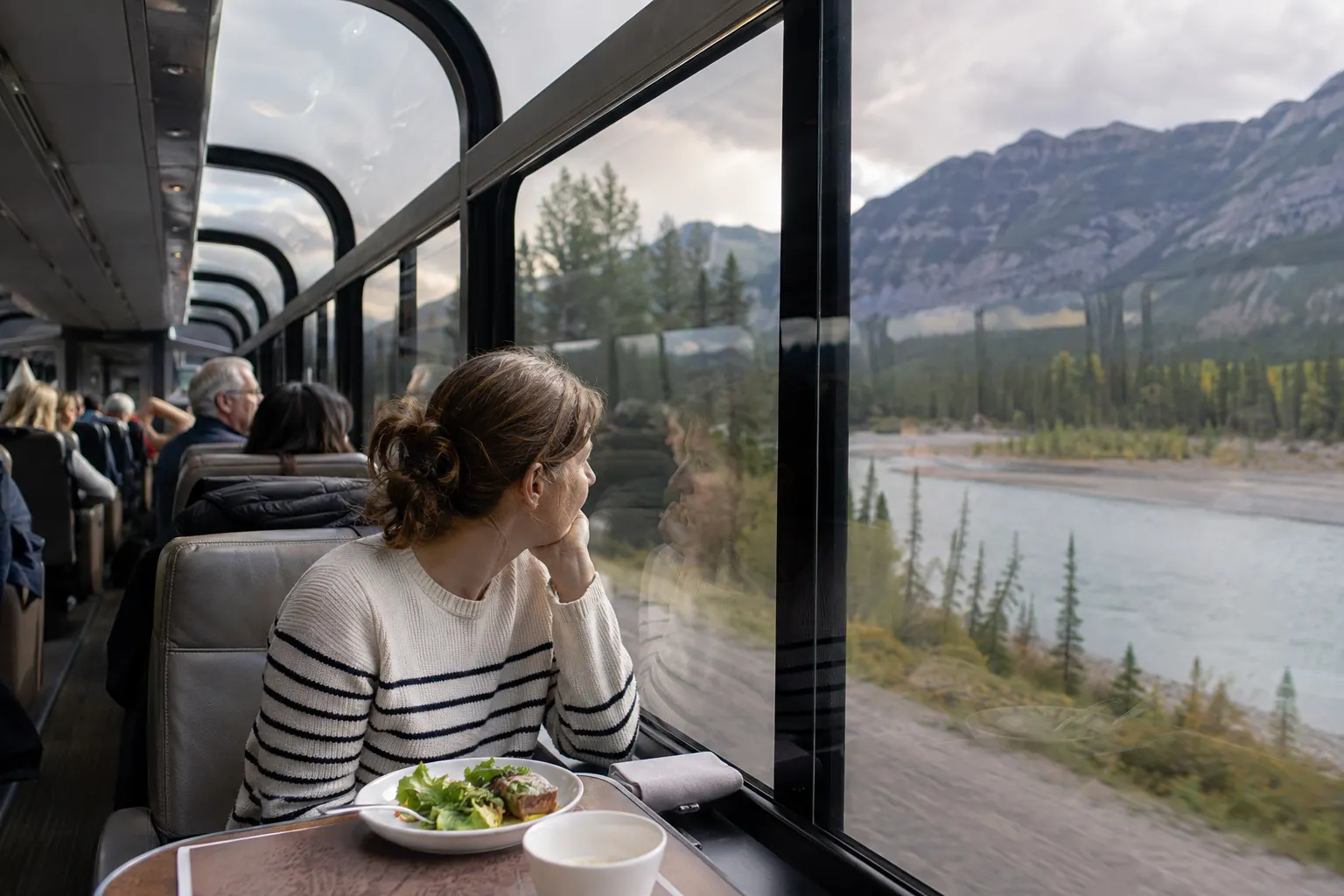 Traveler facing forward in an unbranded panoramic train overlooking a Canadian mountain river valley.