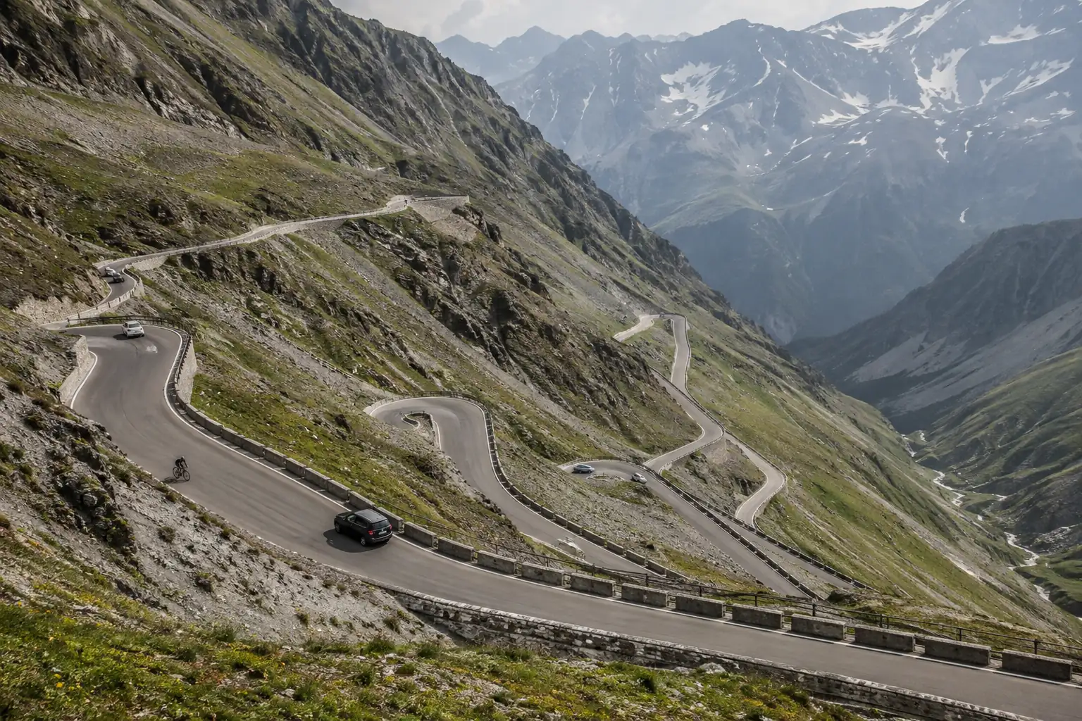 Hairpin bends crossing an alpine slope, with small cars and a cyclist moving within their lanes.