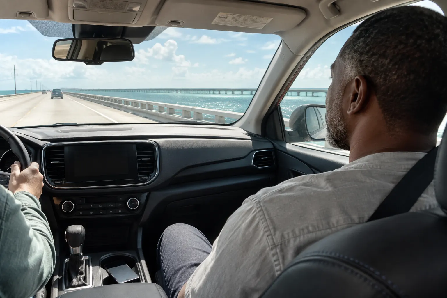 Belted front-seat passenger looking ahead at a bridge and sea horizon during an over-water drive.