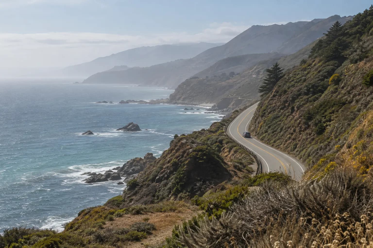 Coastal road curving above the Pacific, seen from a protected overlook through salt haze.