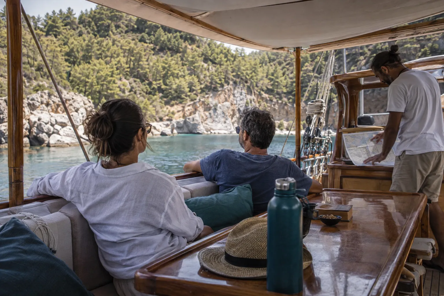 Guests and a crew member on a wooden motor-sailer in a sheltered coastal cove.