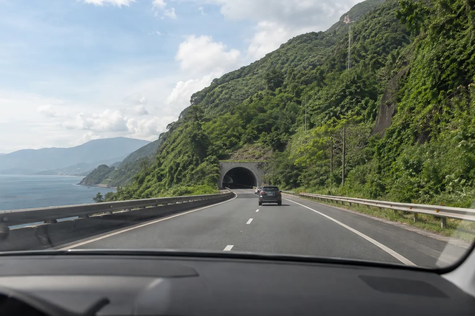 View from a passenger seat toward a broad road tunnel through a green hillside.