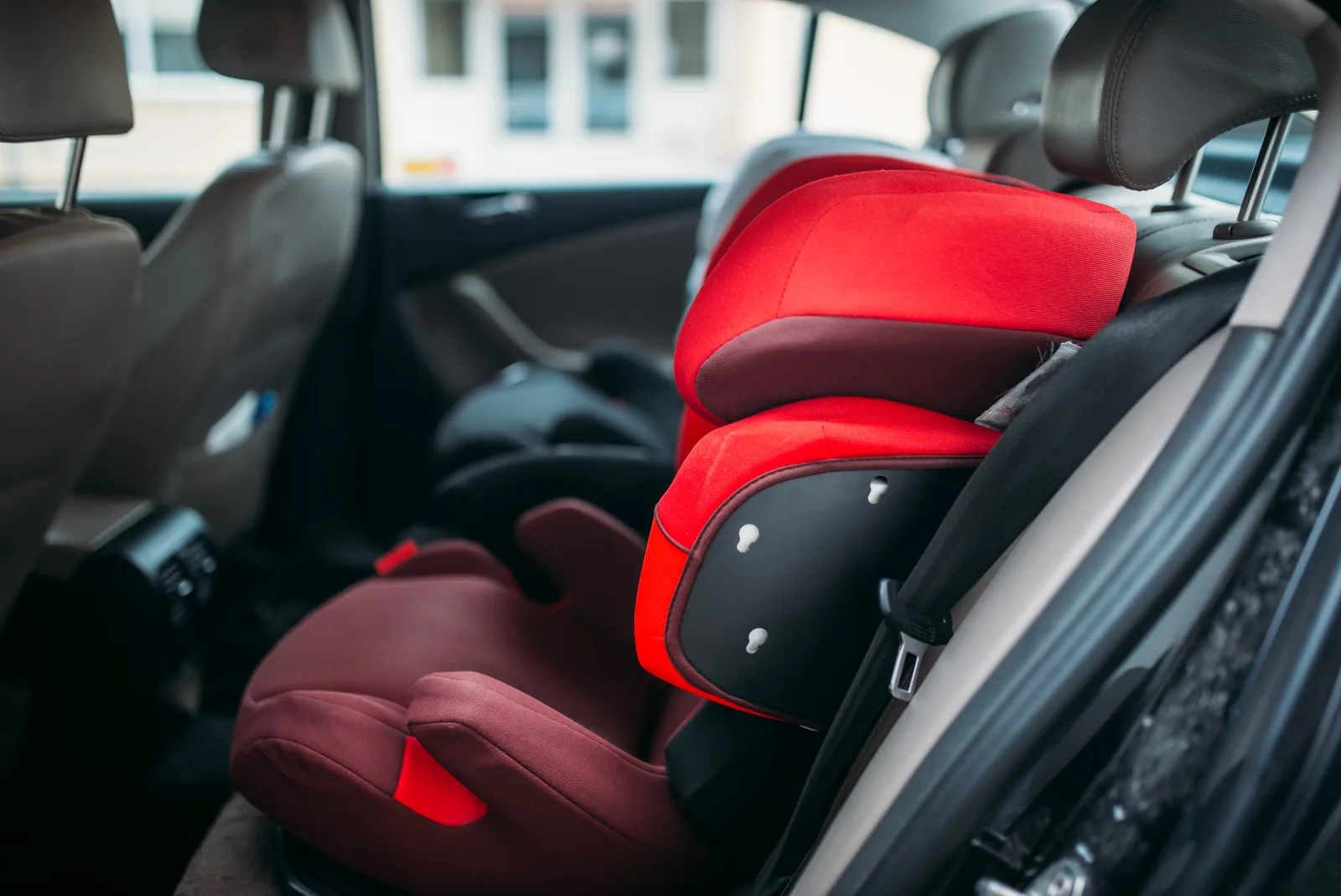 An empty red high-backed child seat strapped into the rear of a car, with daylight coming through the side window.