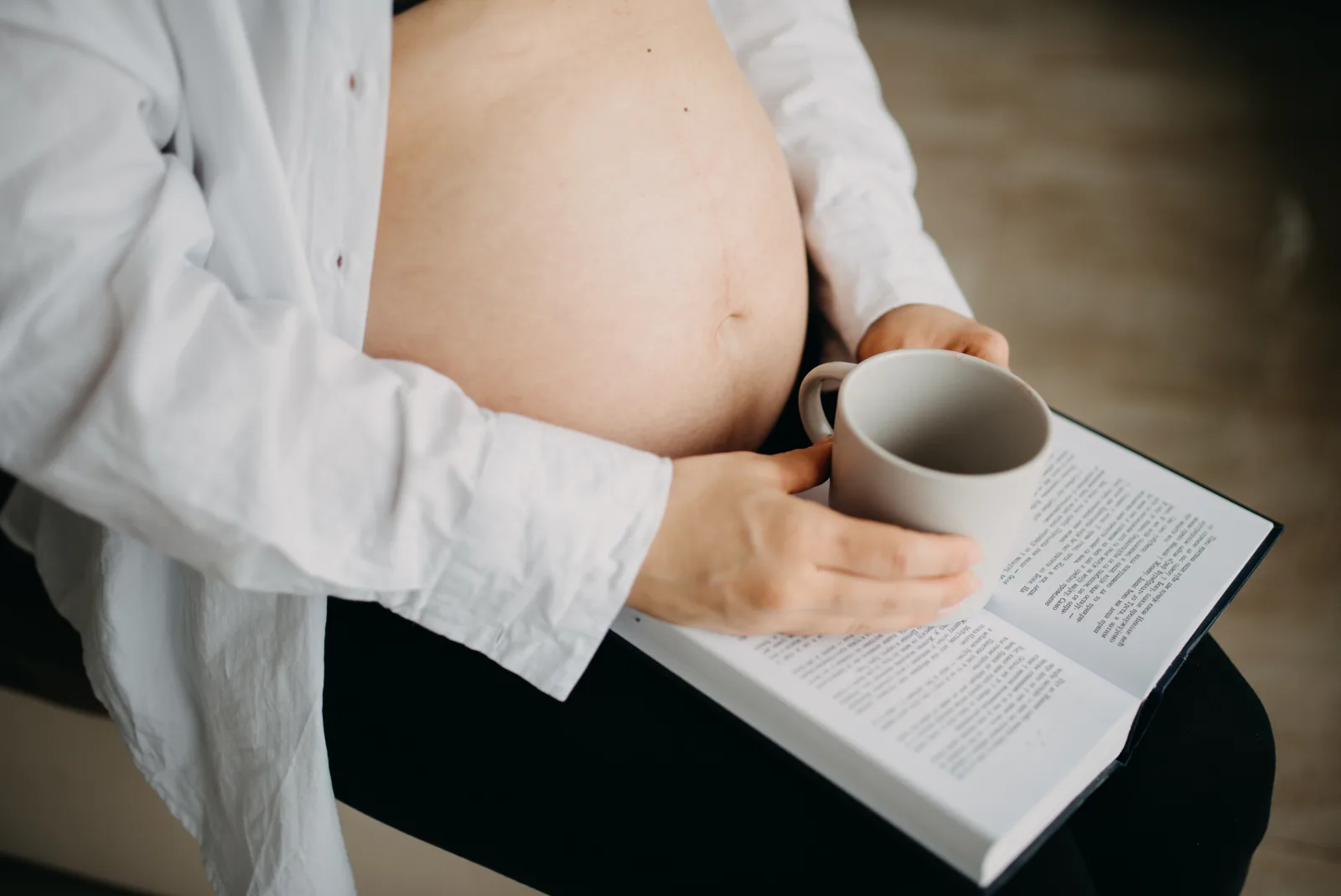 Pregnant woman seated at home with an open book on her lap, holding a mug in front of her bare belly in soft natural light.