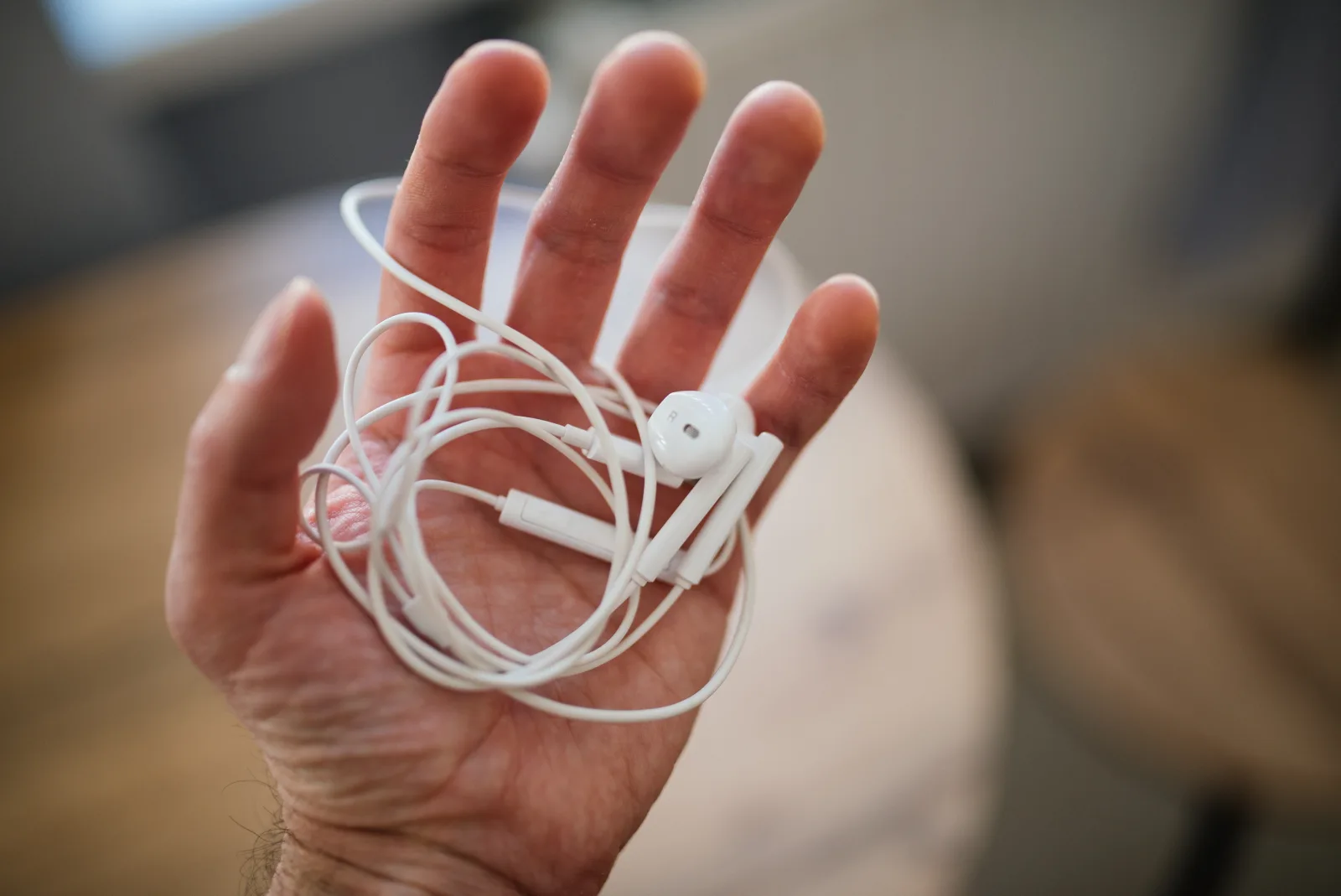 An open hand holding a pair of white wired earbuds in a bright room with a blurred wooden table behind.