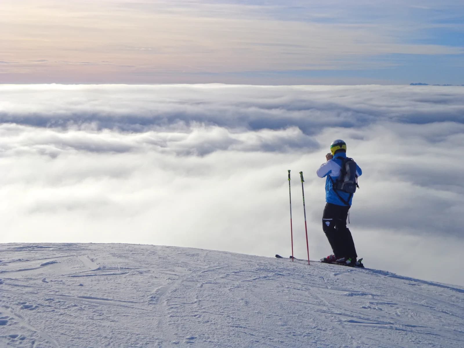 A skier standing still at the edge of a groomed piste, seen from behind, looking out over an unbroken sea of cloud that hides the horizon.