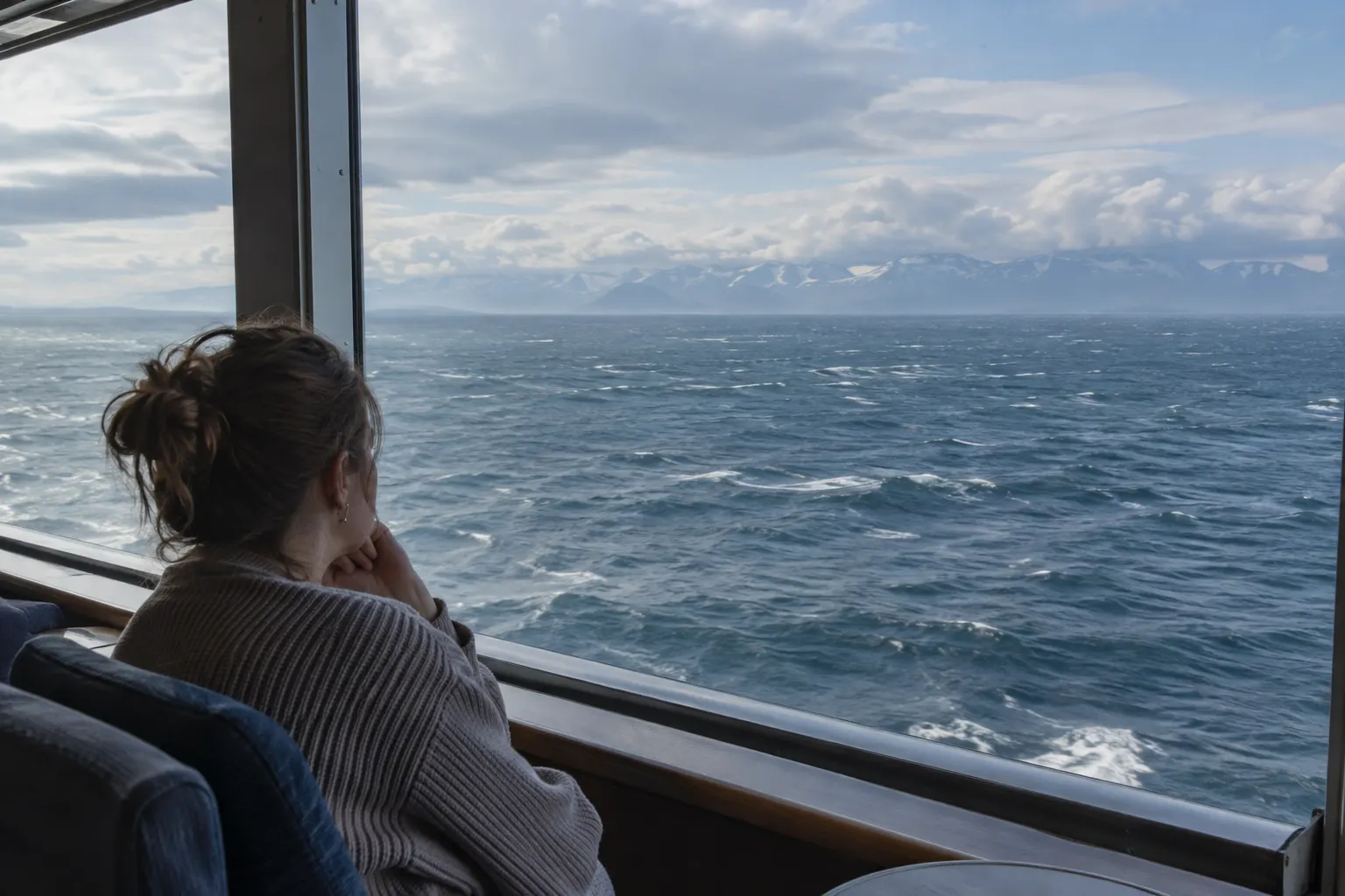 Open sea with distant snow-streaked coastal mountains viewed from a ship's observation area.