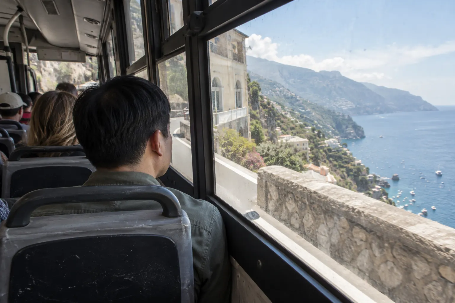Bus passenger looking through the window at a coastal road bend, with the sea beyond.
