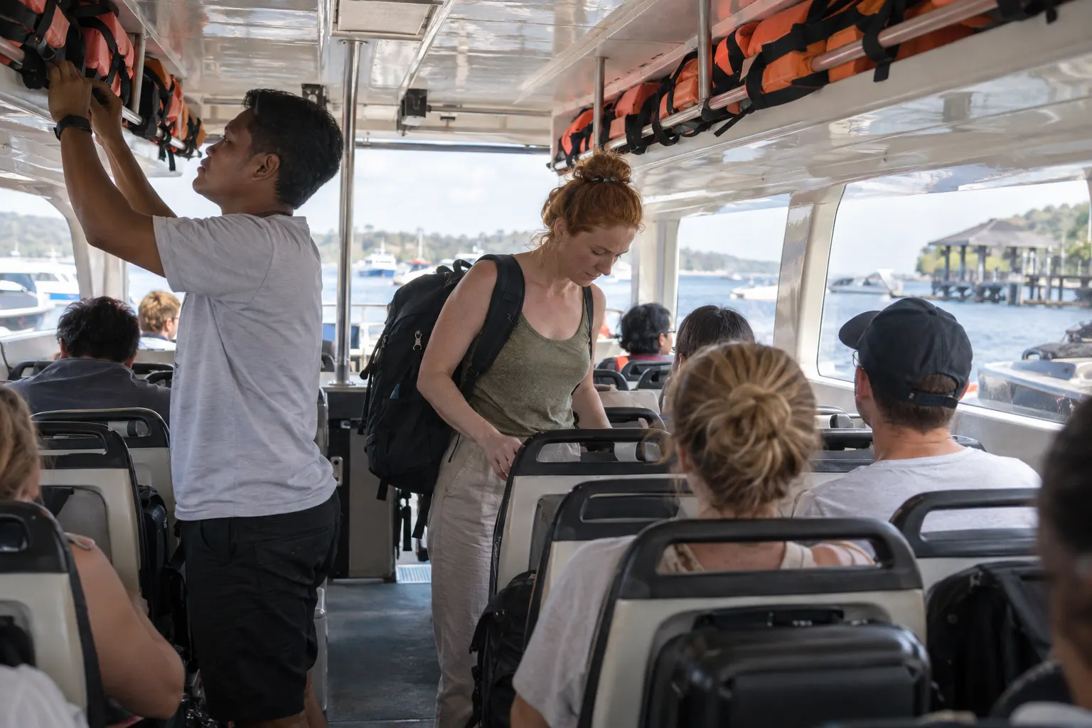 Passengers settling into a fast-boat cabin while a crew member checks secured life jackets.