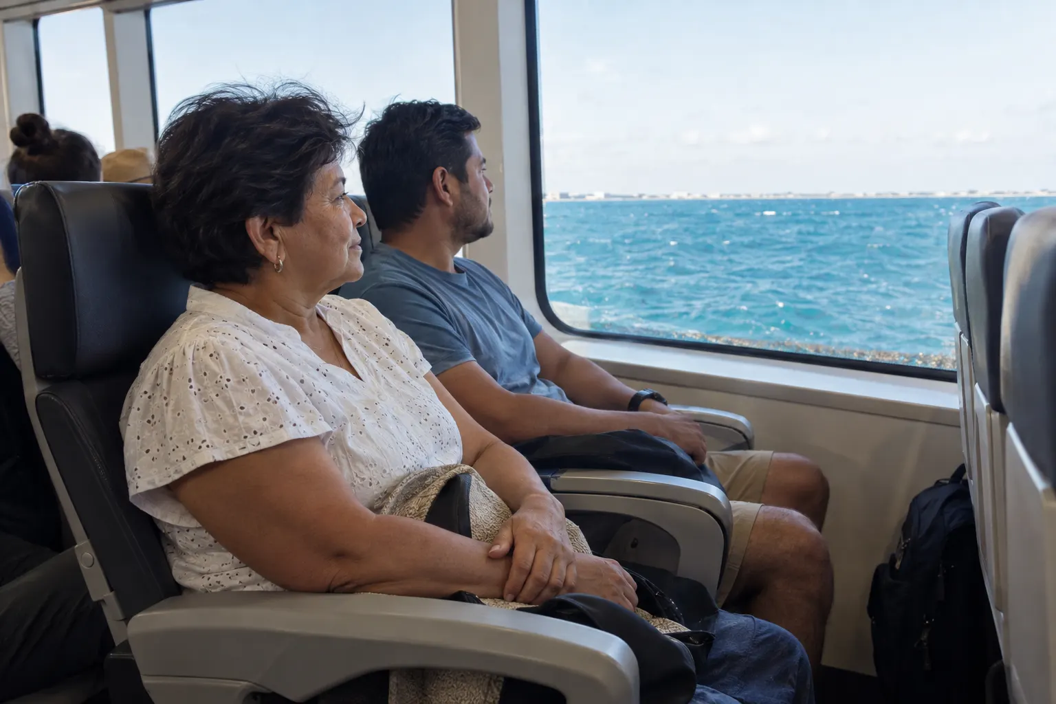 Two ferry passengers watching turquoise channel water from seats in the middle of the cabin.
