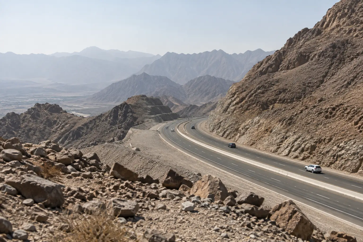 Road bending between bare rocky mountain slopes under pale daylight, with distant cars.