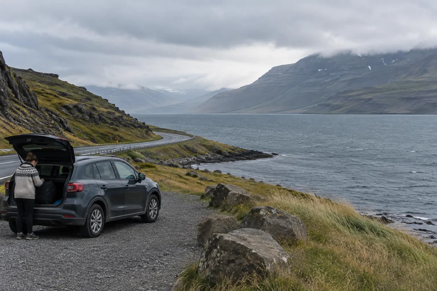 Road following a fjord beneath low cloud, viewed from a safe pullout with a parked car.