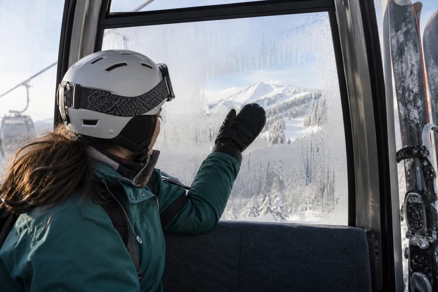 Seated skier clearing condensation from a gondola window to see the distant ridge.