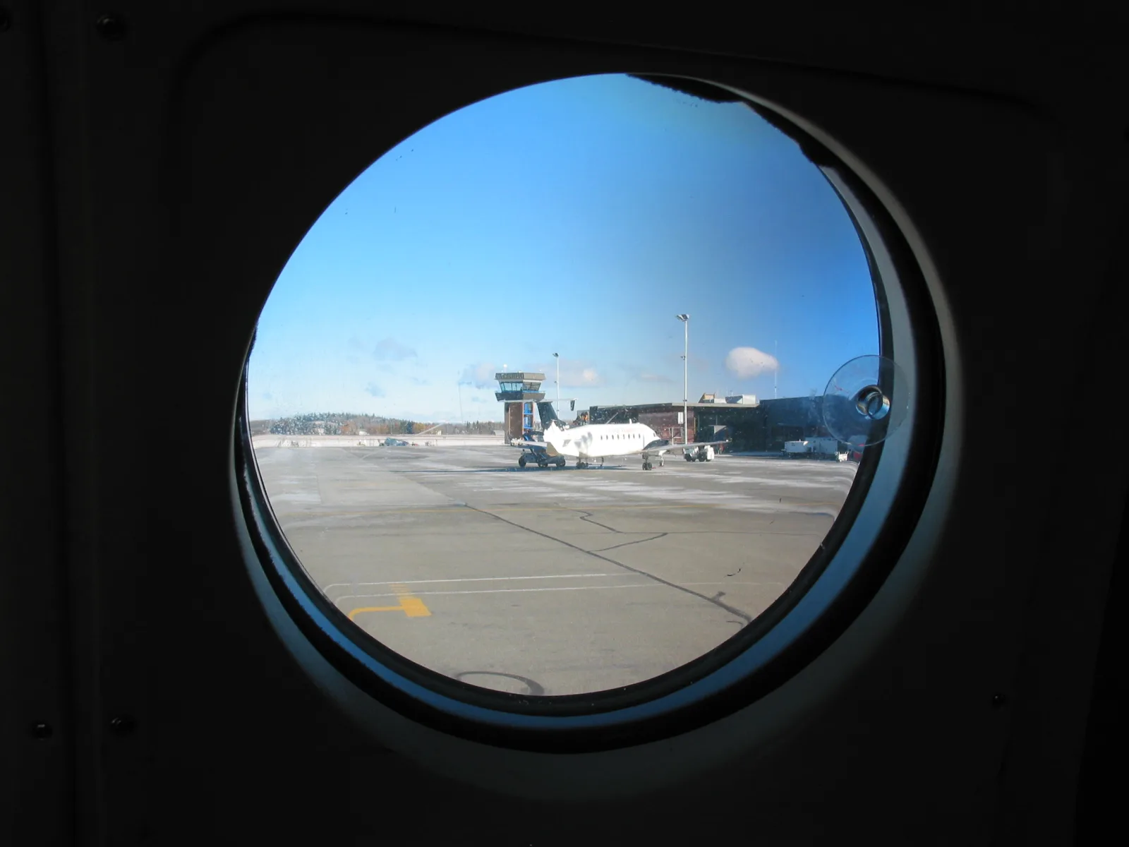 A round aircraft cabin window looking out onto an airport apron, where a small white turboprop stands parked in front of a control tower under a clear blue sky.