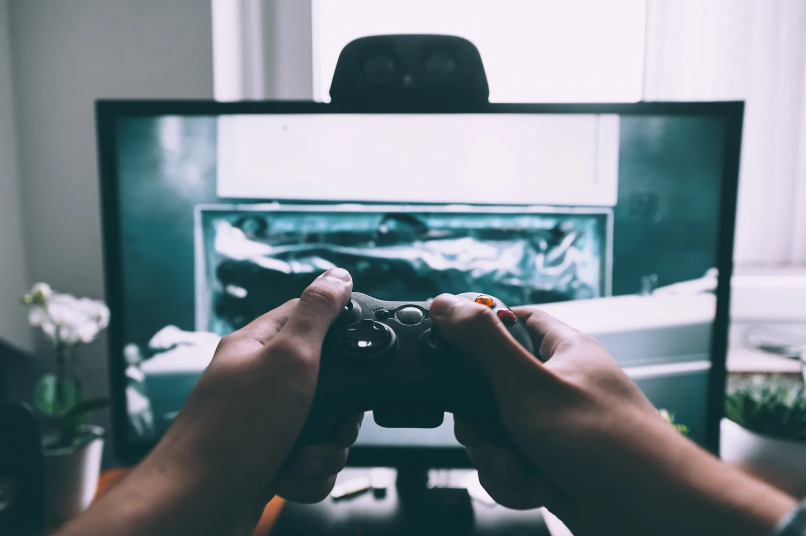 Player's hands holding a game controller in front of a large monitor in a dim room lit by the screen.