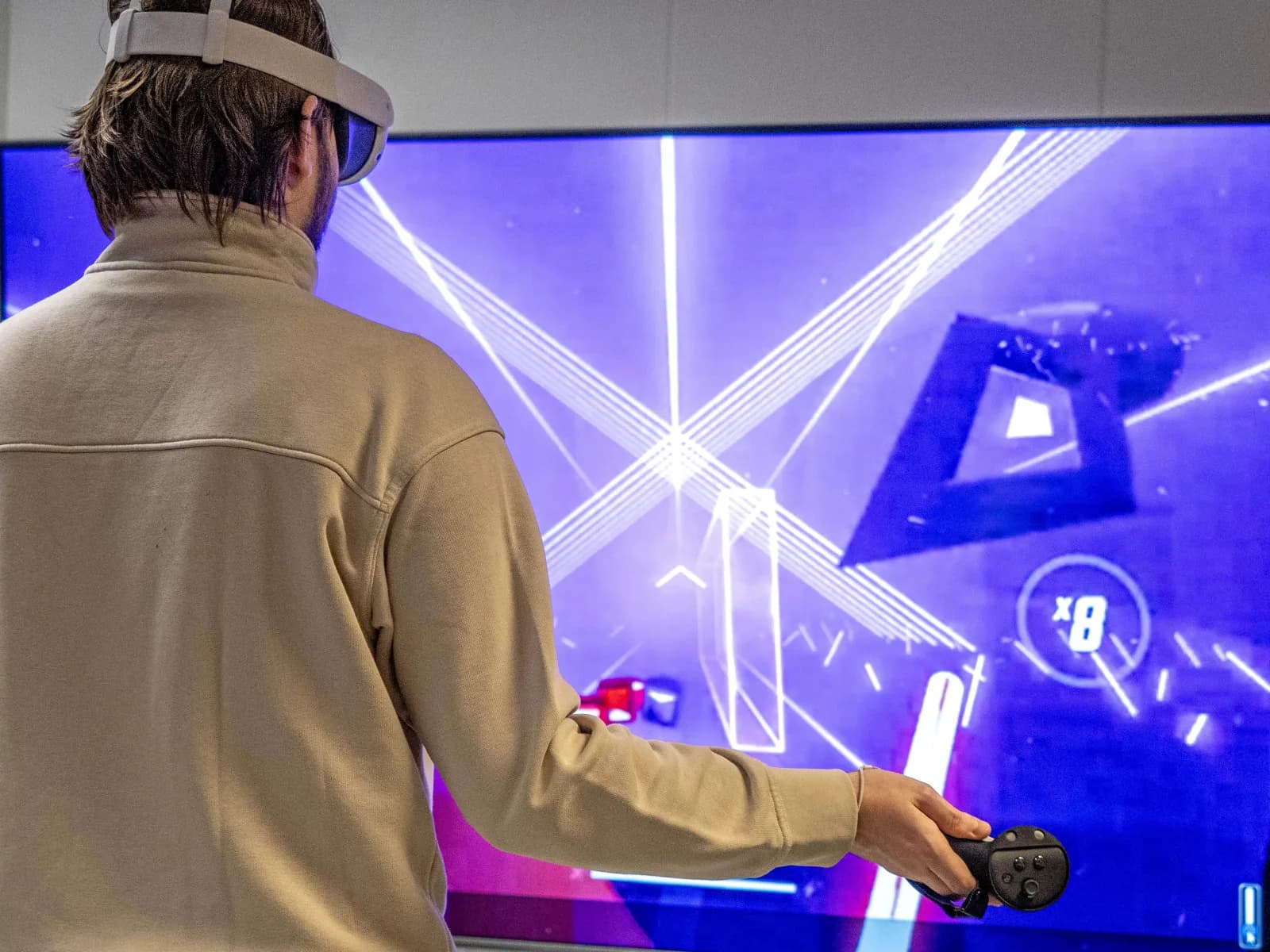 A person seen from behind wearing a VR headset and holding a motion controller in front of a large screen mirroring the game.